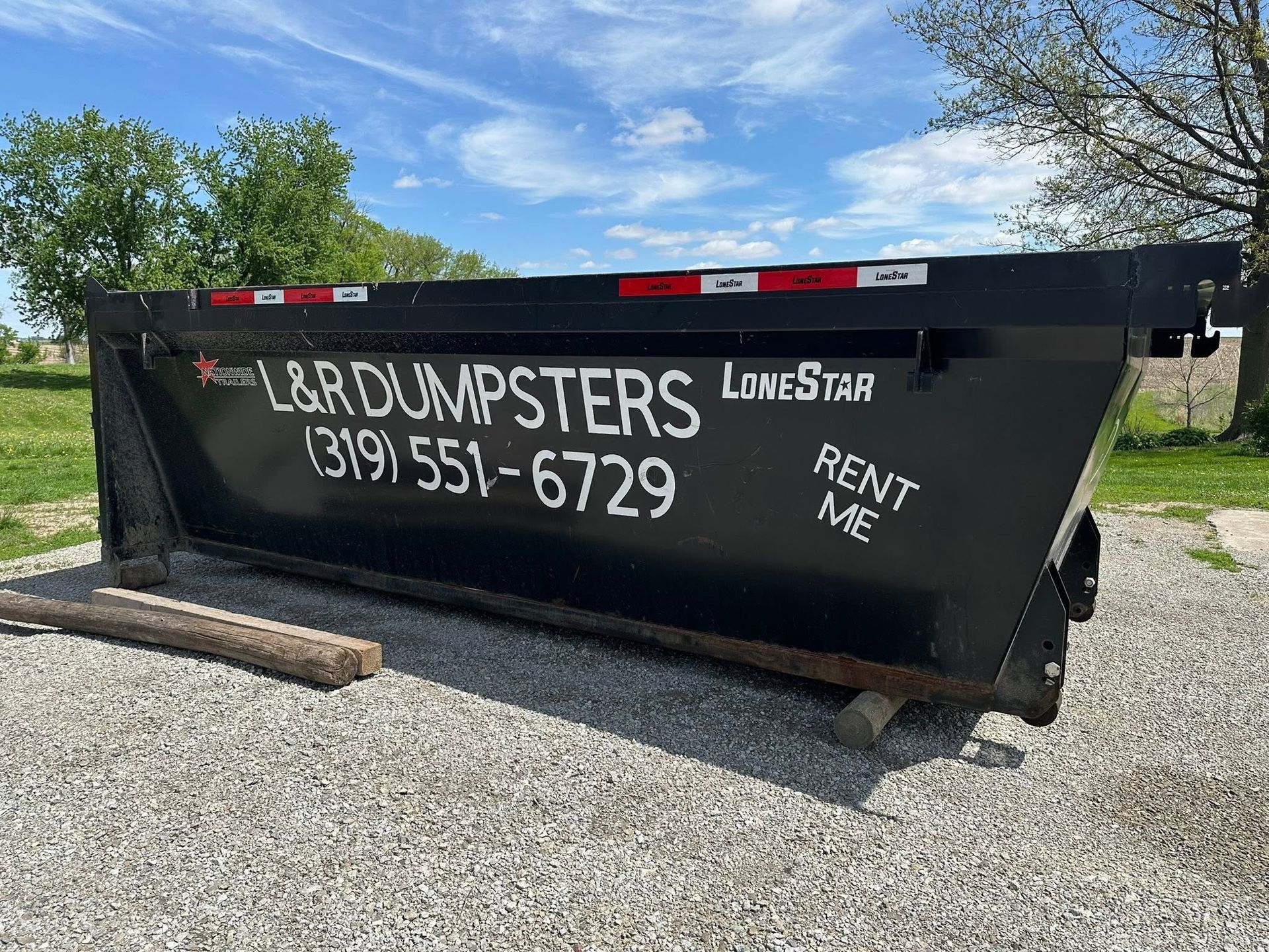 Black dump trailer with “L&R Dumpsters” and phone number on gravel lot under blue sky