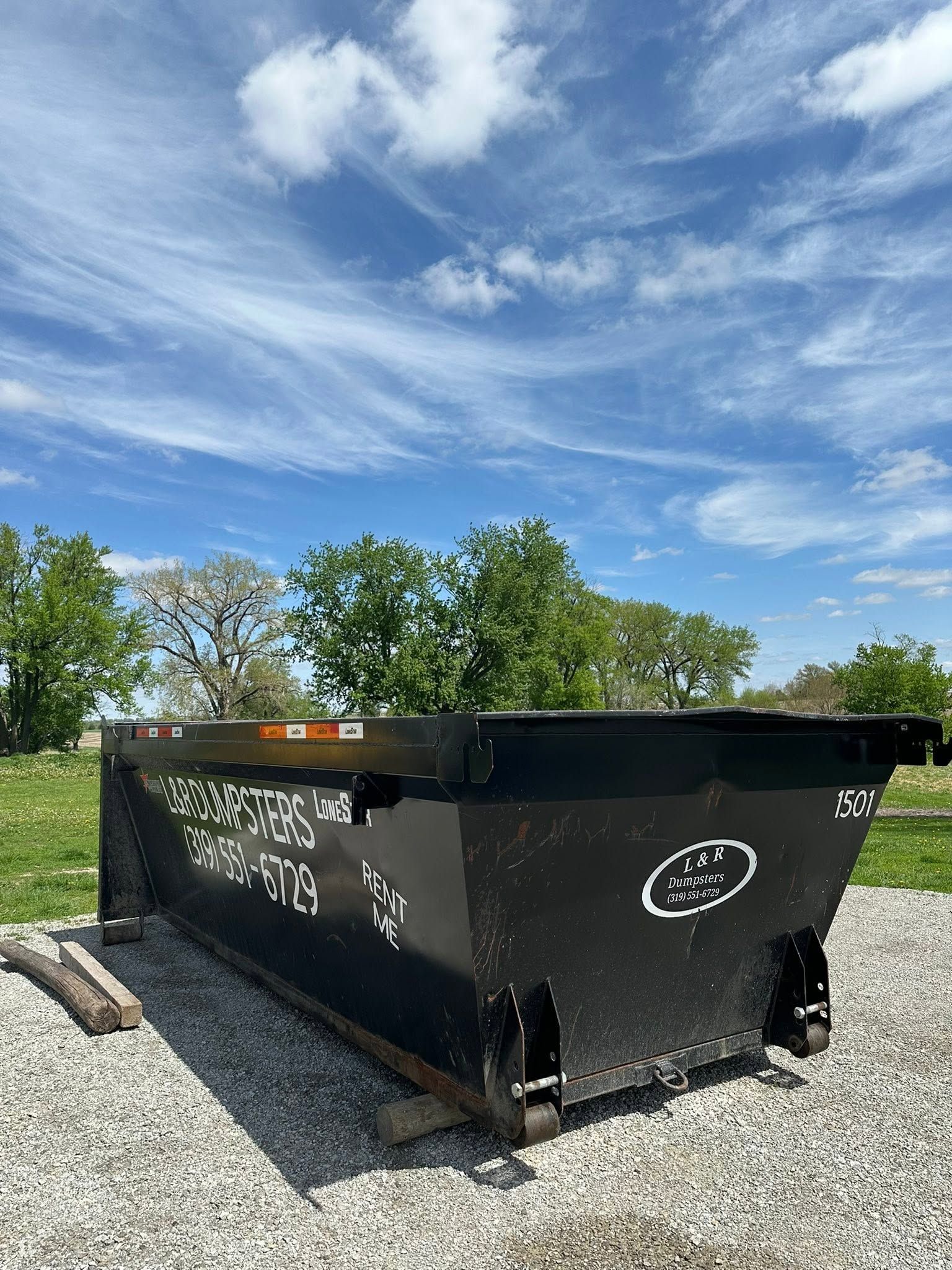 Black dumpster on gravel lot with trees and blue sky in the background