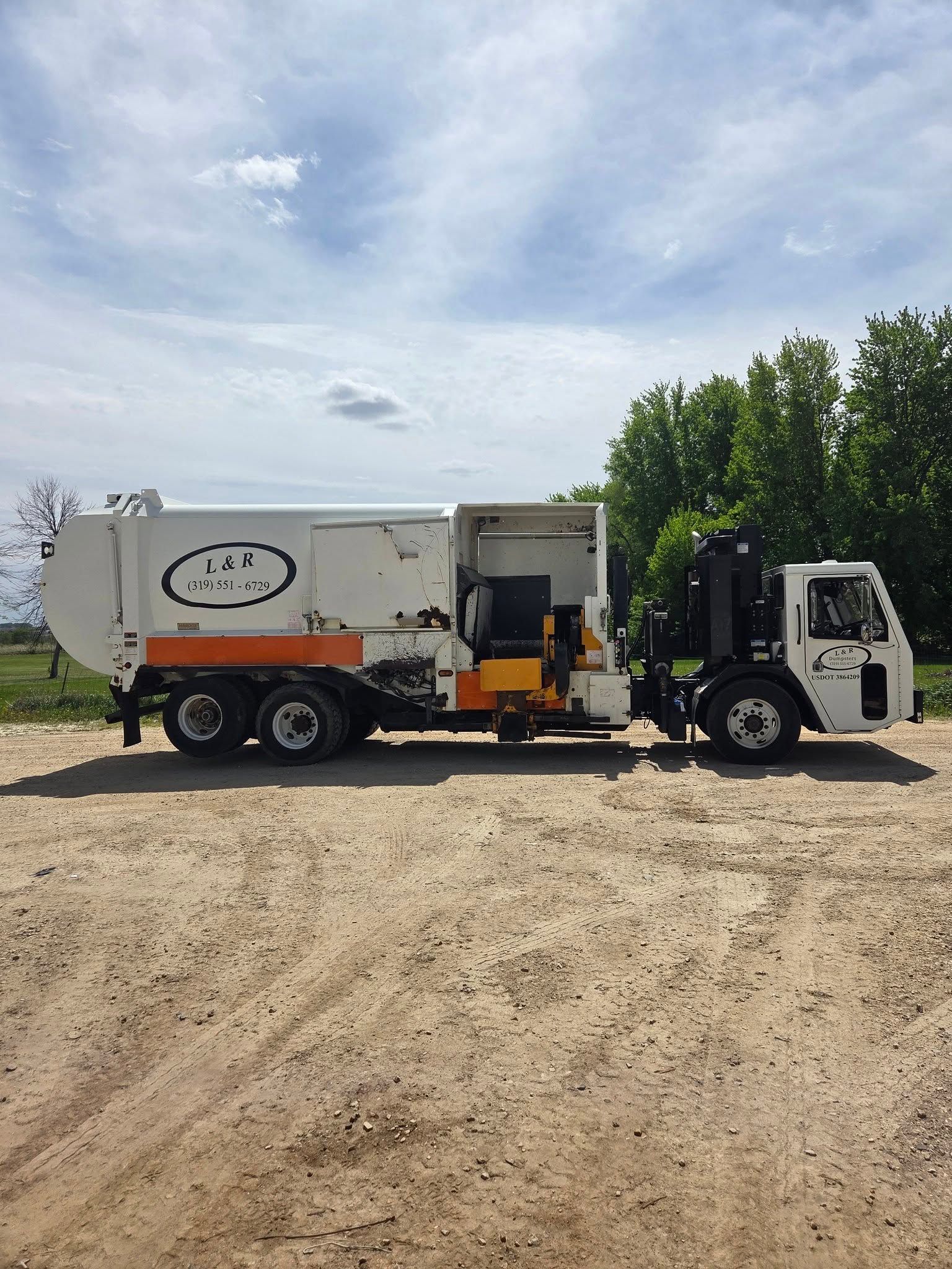 Front loader truck parked on gravel, side view, with open rear compartment under a cloudy sky.