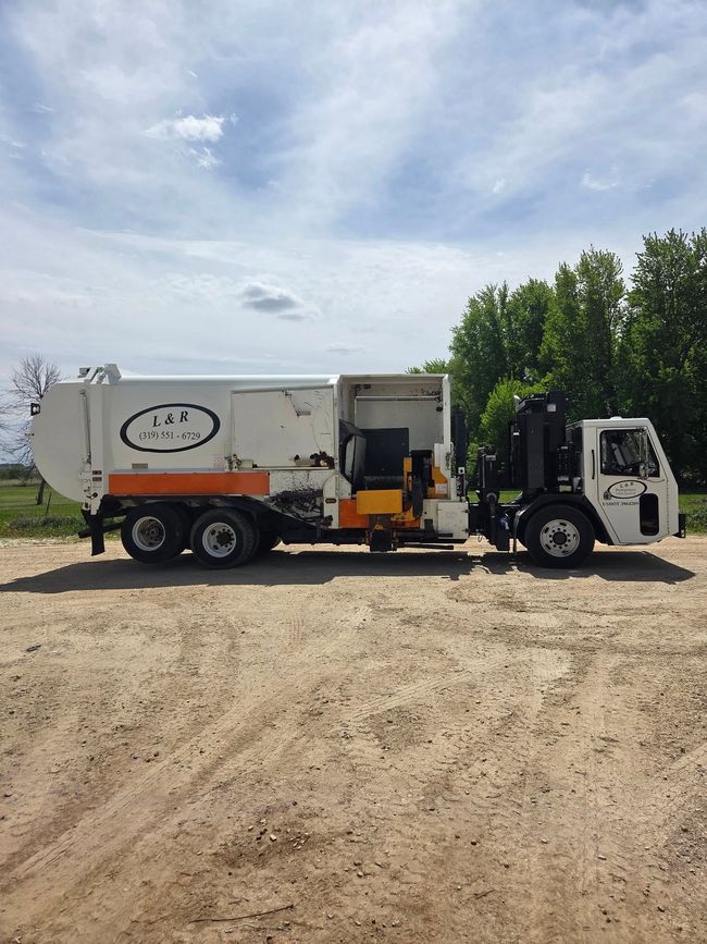 White garbage truck with side loader parked on a dirt lot under a cloudy sky