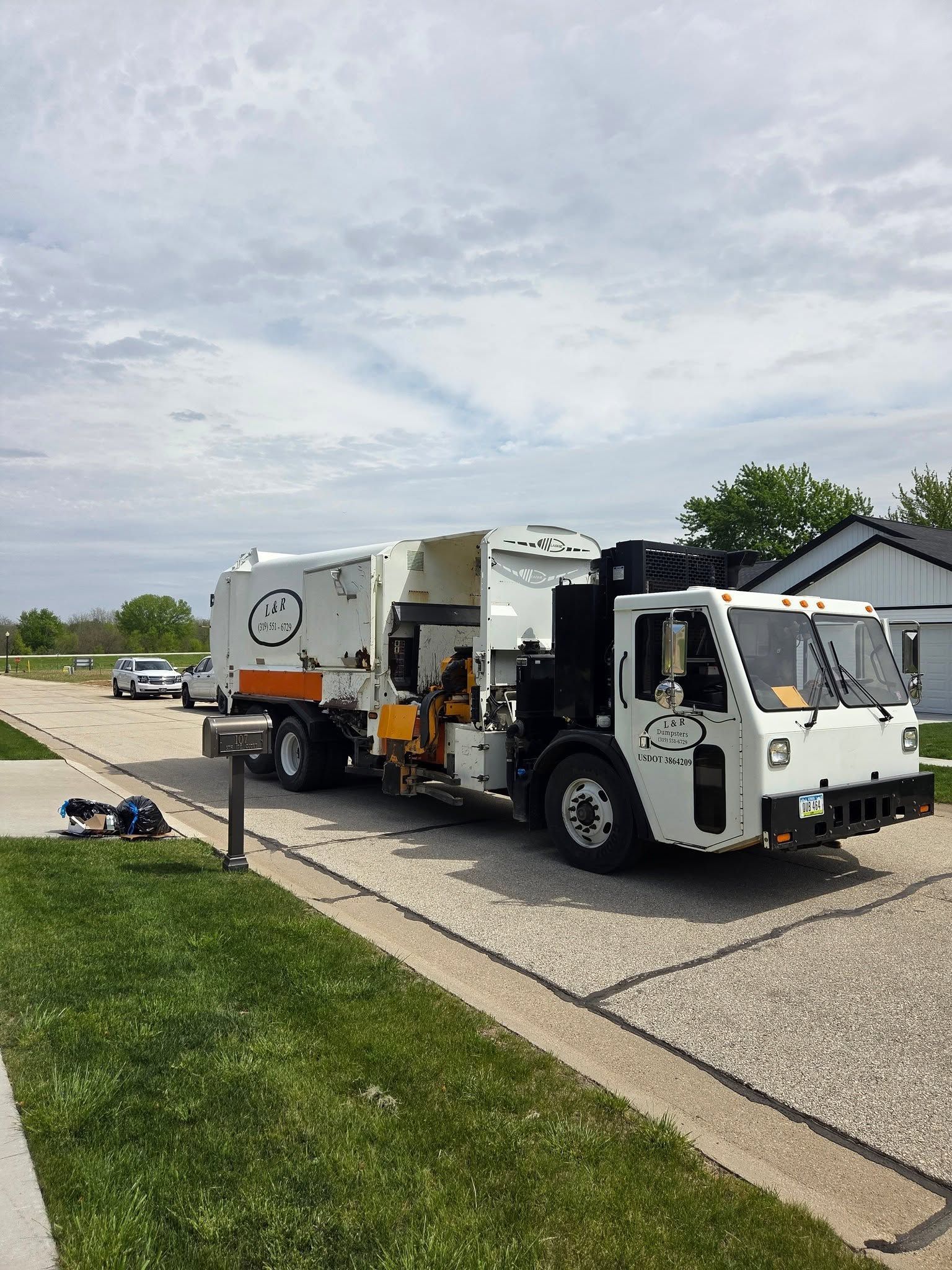 White garbage truck parked on a suburban street beside a driveway under a cloudy sky