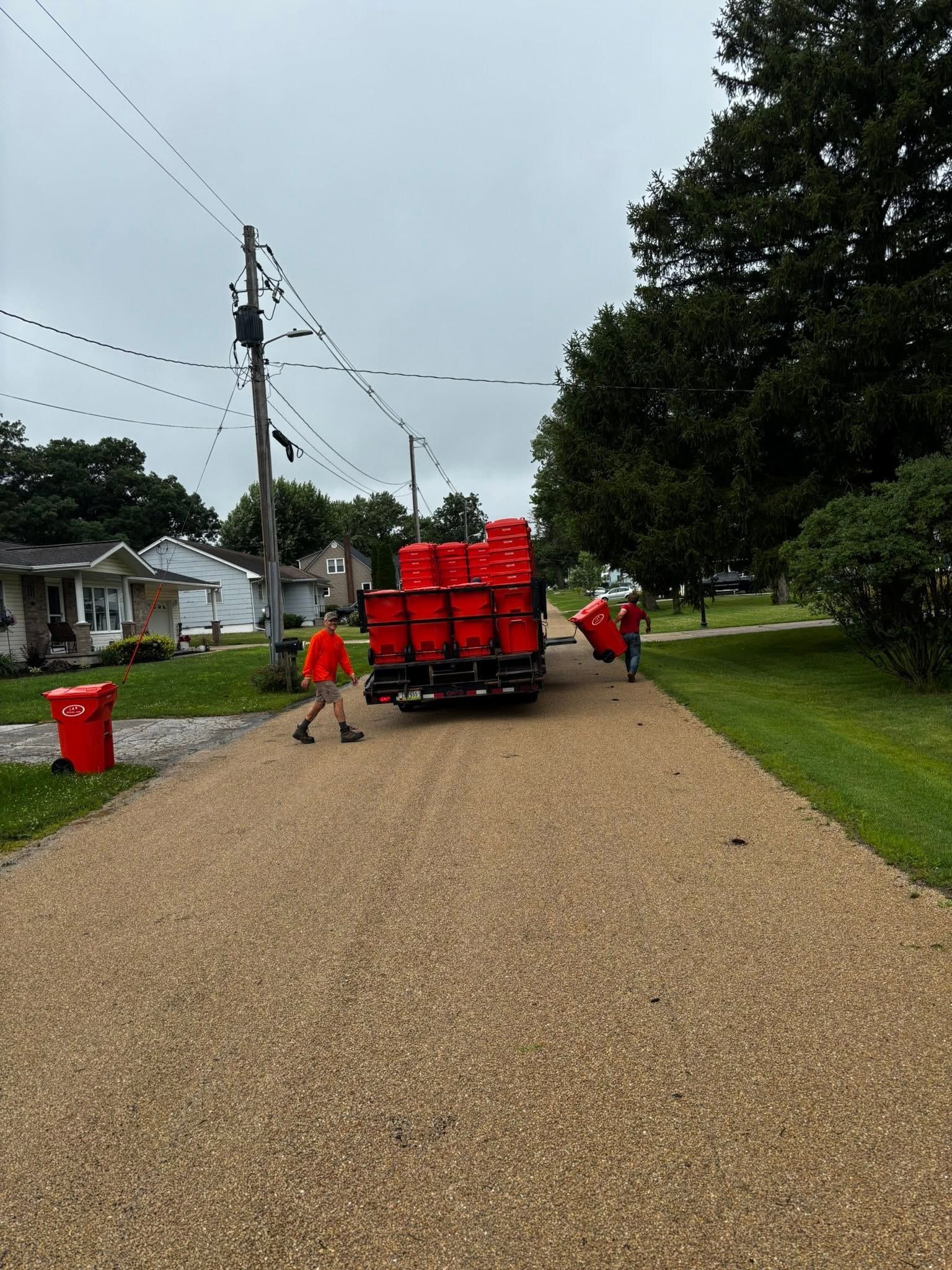 People loading bright red bins onto a truck on a gravel neighborhood road under cloudy skies