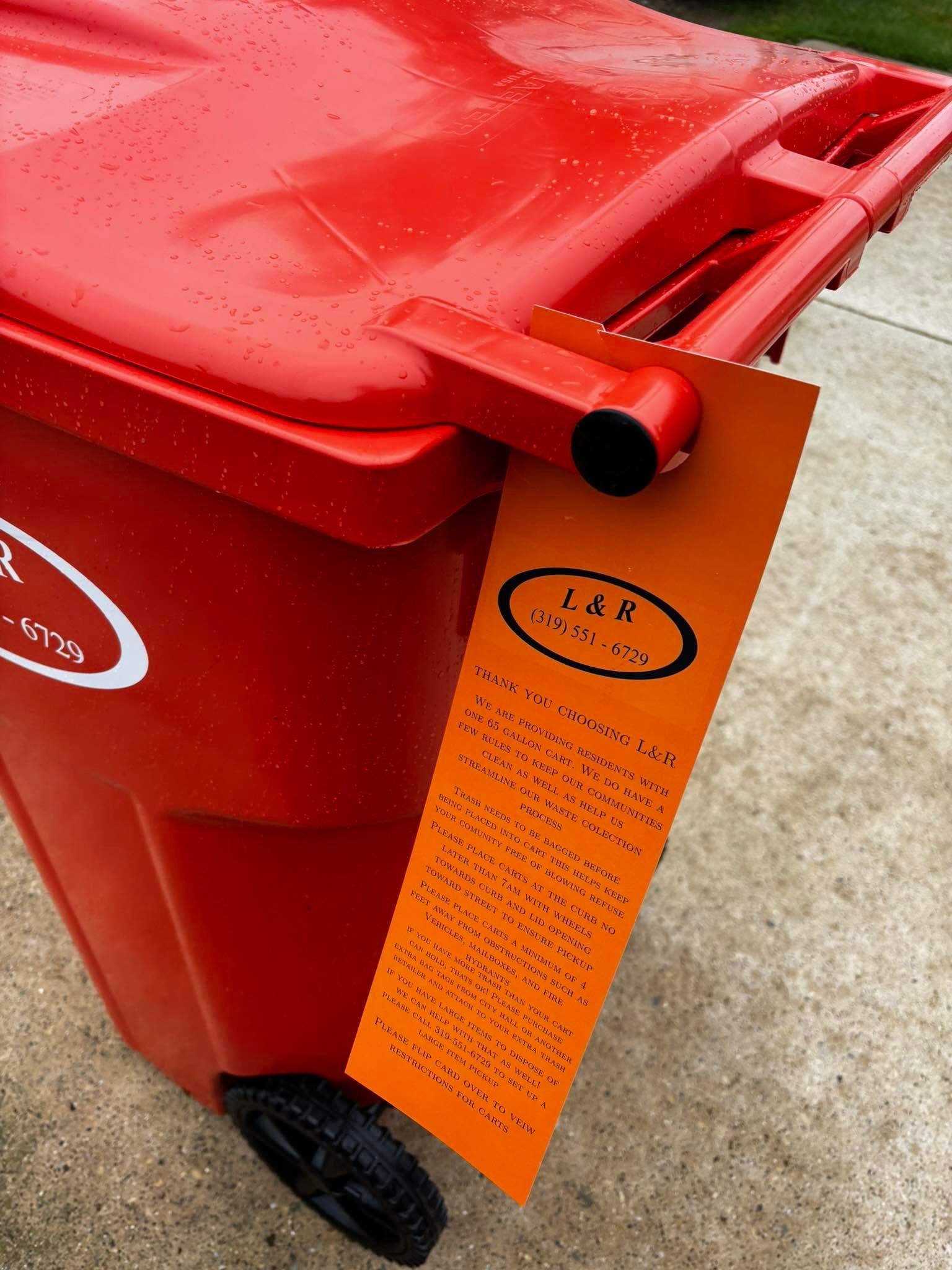 Red wheeled trash bin with an orange information tag hanging from the lid handle.