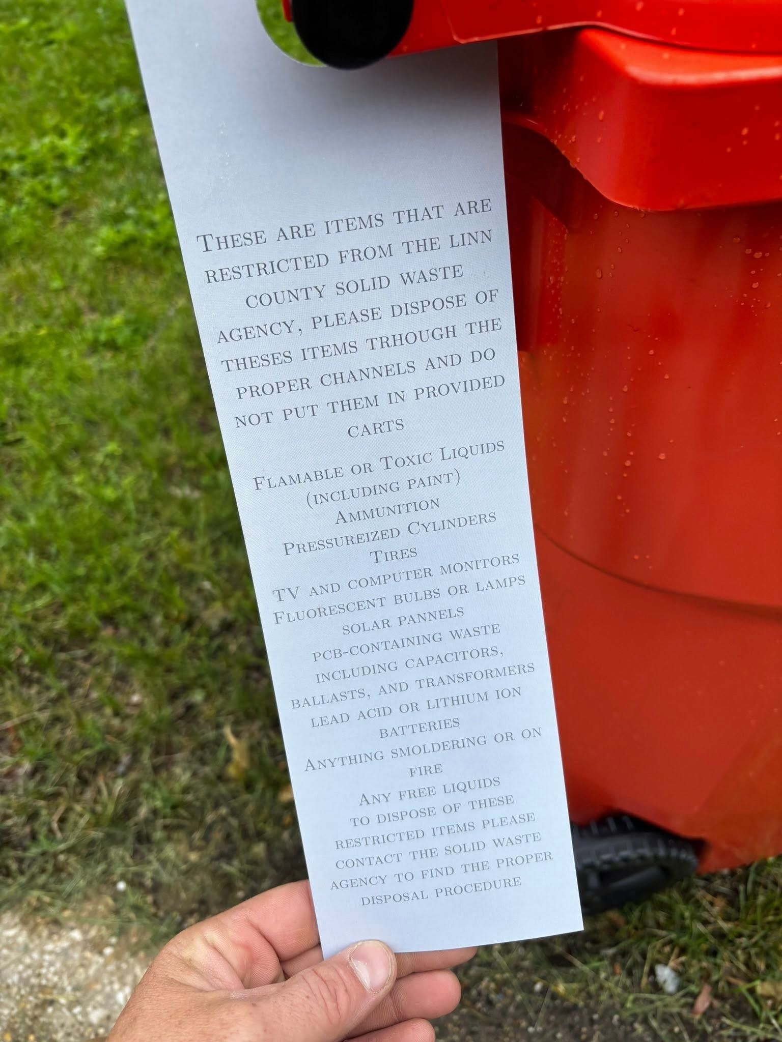 Hand holding a long receipt beside a red trash bin outdoors on grass.