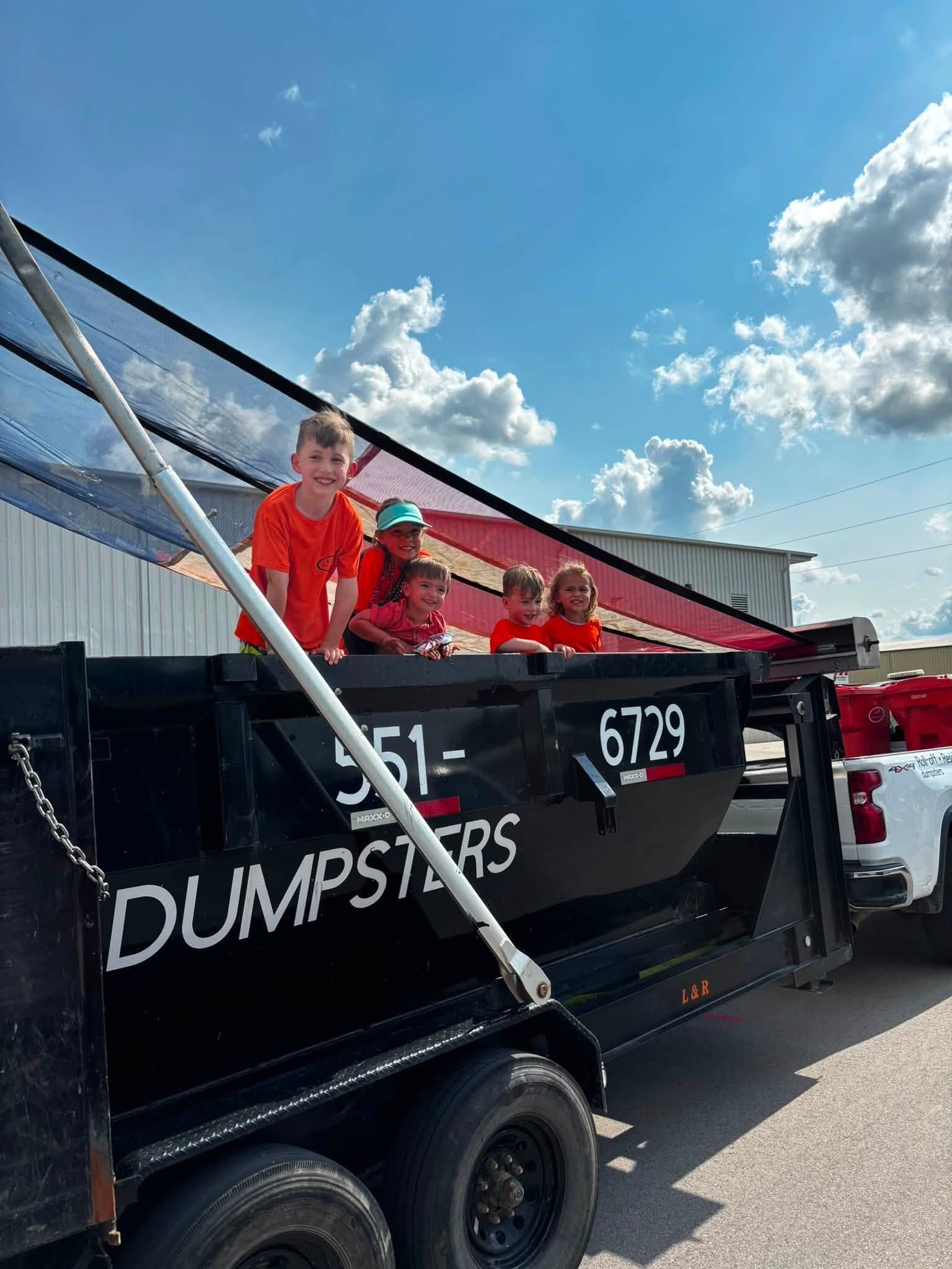 Workers in orange vests sit in a black dumpster trailer under a blue sky.
