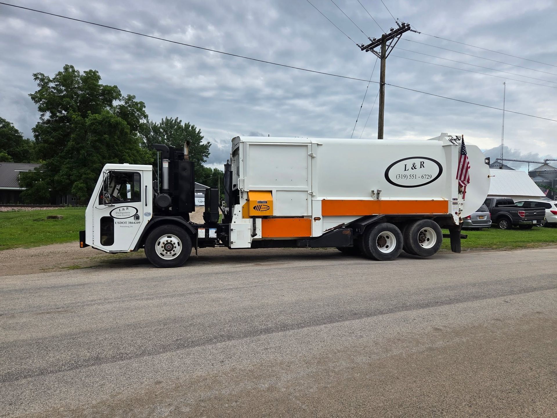 White-and-orange garbage truck parked on a gravel roadside under cloudy skies