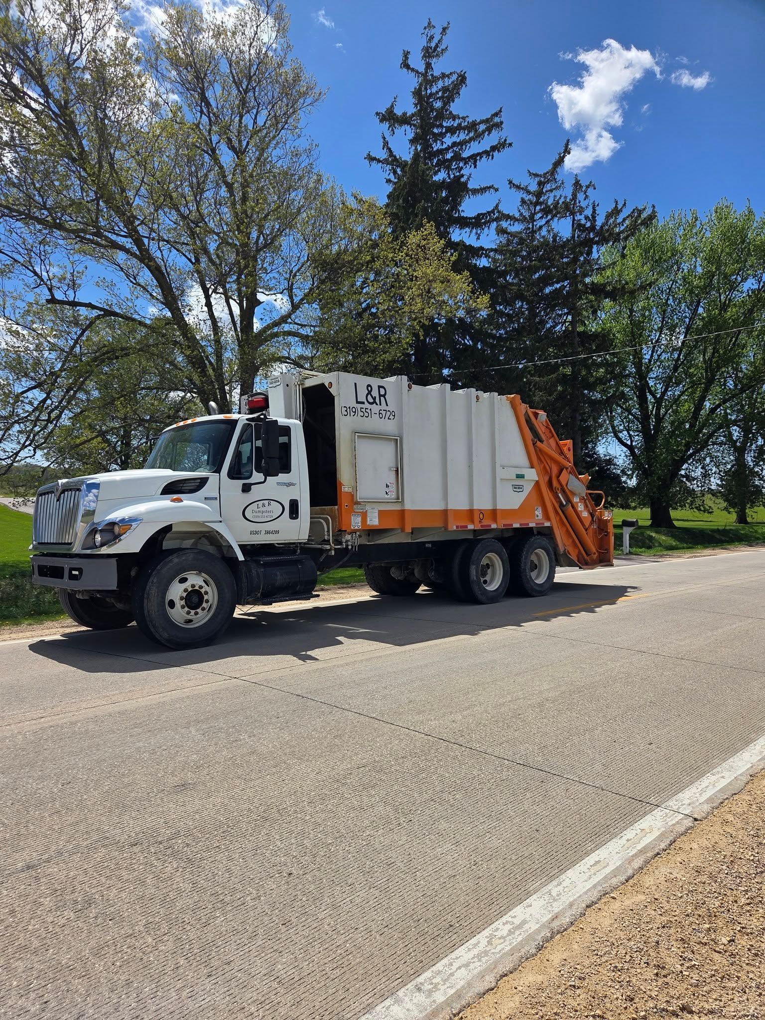 White garbage truck on a sunny road beside trees and blue sky