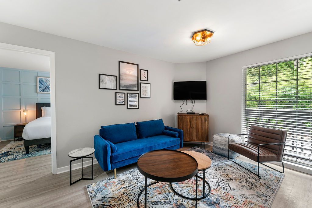 Bright living room with blue sofa, round coffee table, wall TV, and large window with blinds.