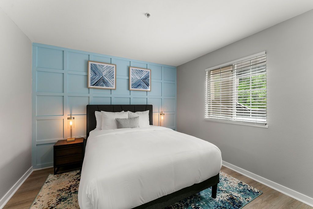 Bright bedroom with a bed, blue accent wall, bedside lamps, and a window with blinds