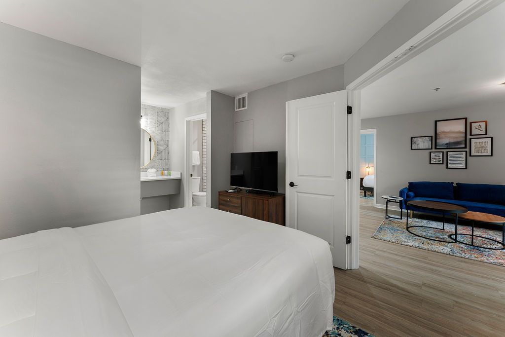 Bedroom with white bed, dresser, TV, and open door to a sitting area with blue sofa and wall art