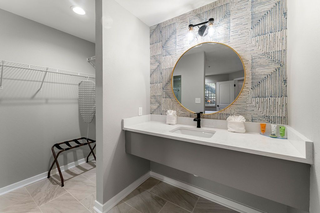 Modern bathroom vanity with round mirror, white countertop, and adjacent open closet area.