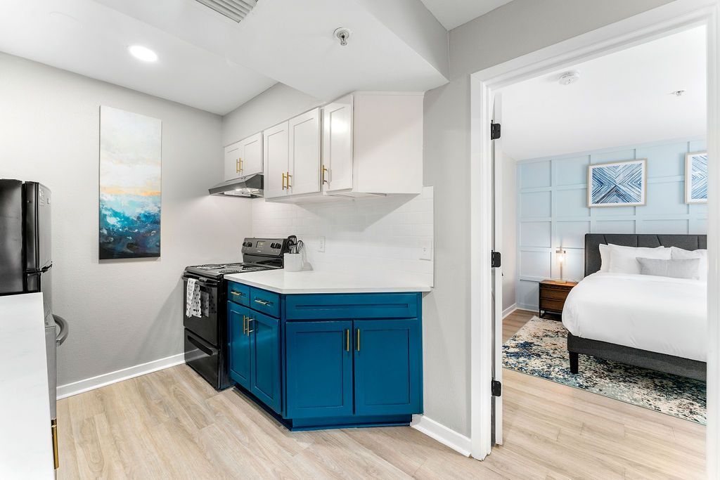 Bright kitchen with blue cabinets and a doorway leading to a white bedroom with a bed and framed art.