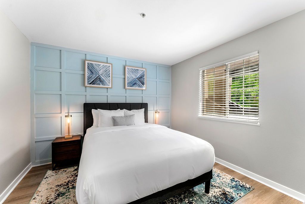 Bright bedroom with a white bed, pale blue wall panels, nightstands, and a window with blinds.