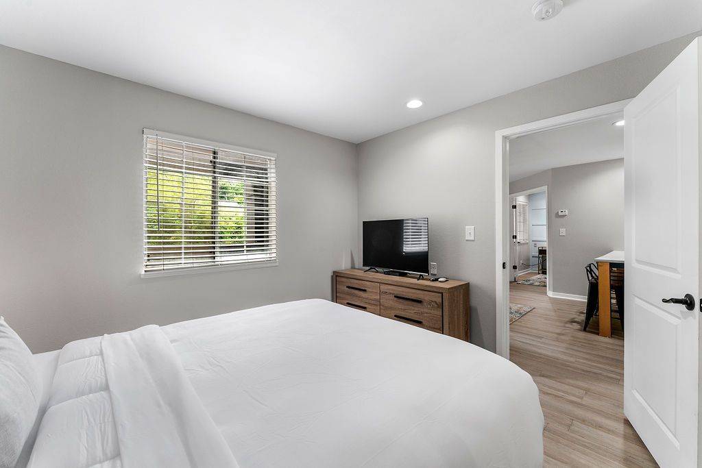 Bright bedroom with a white bed, gray walls, dresser, TV, and an open door to a hallway