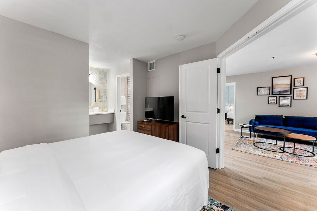Bright bedroom with white bed, open door, TV, and glimpse of a blue sofa in adjacent room