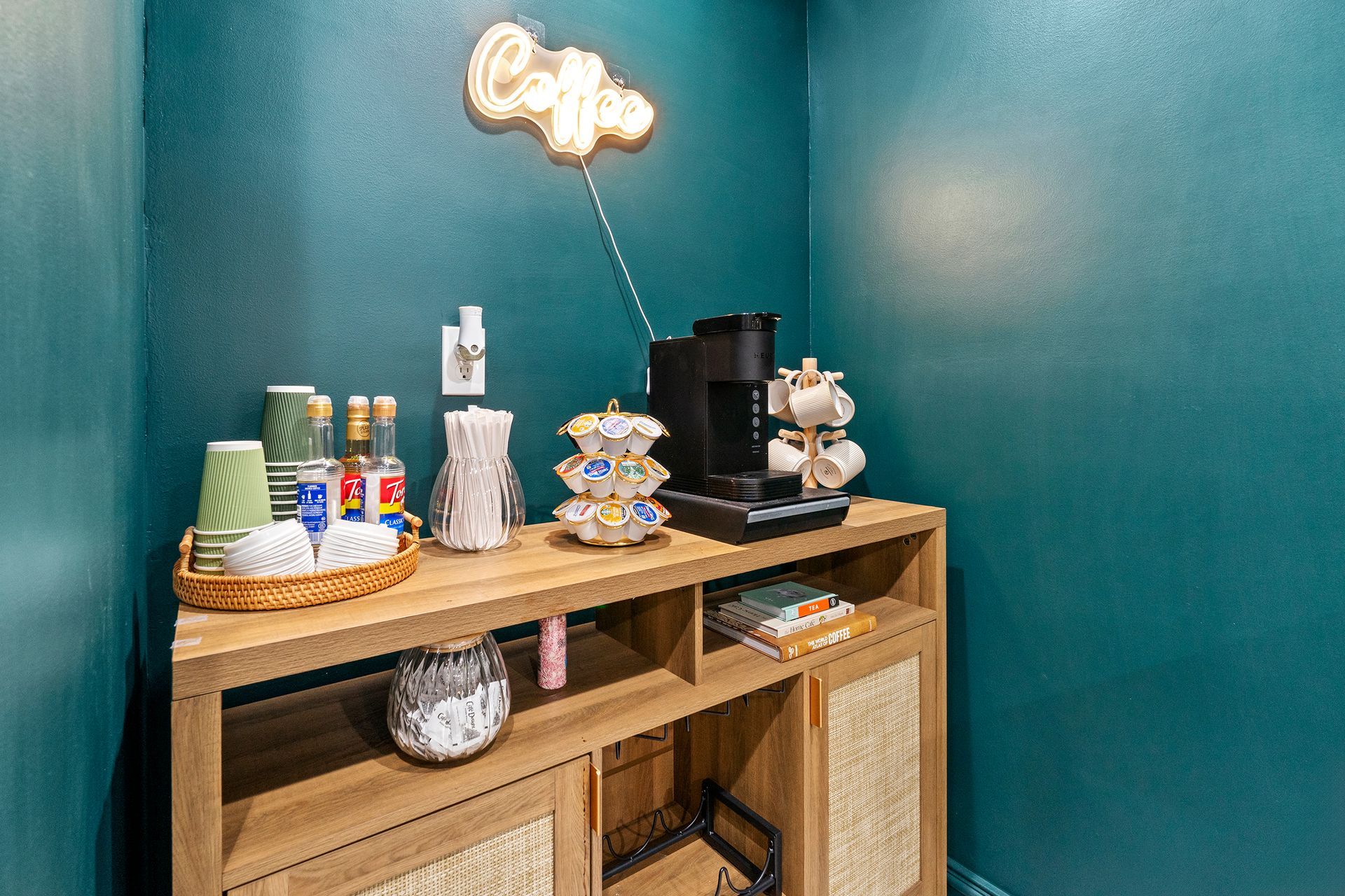 Coffee bar with wooden console, colorful cups, espresso machine, and glowing wall sign against teal walls