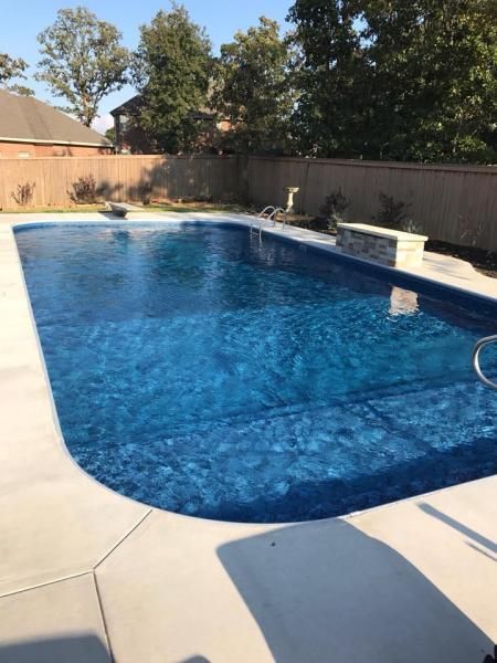 A rectangular swimming pool with deep blue water, surrounded by light-colored concrete decking and a wooden fence.