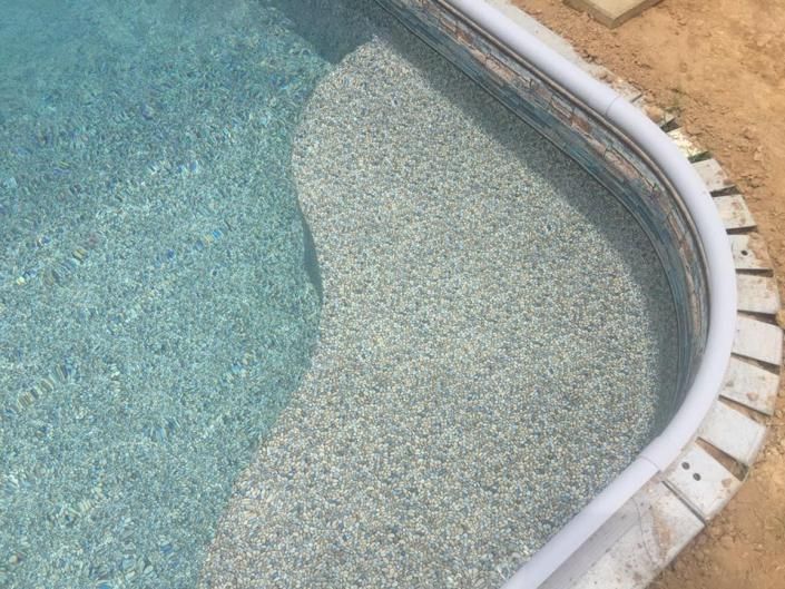 A swimming pool corner featuring a curved entry step with pebble-textured finish and white coping against dirt ground.