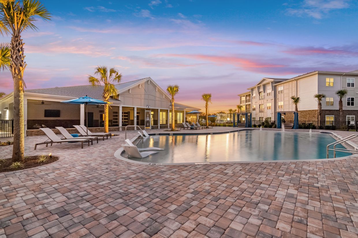 A large swimming pool surrounded by chairs and palm trees in front of a building.