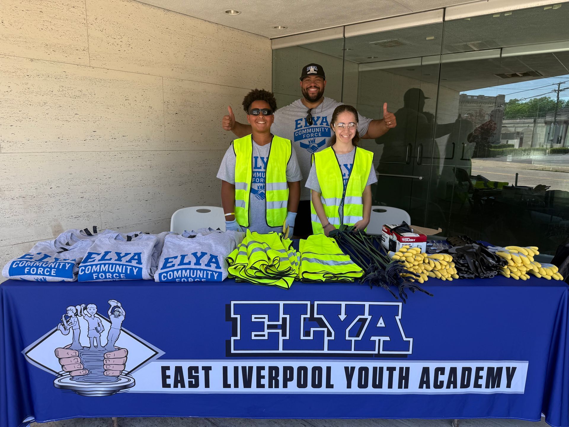 Three volunteers behind a table with ELYA East Liverpool Youth Academy shirts and folded clothing outdoors