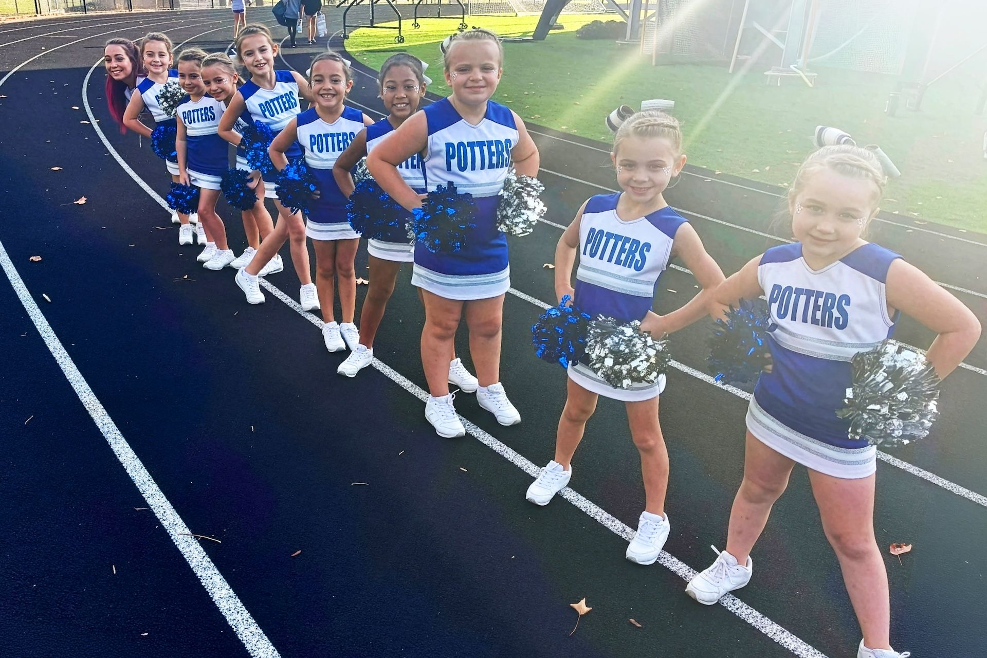 Youth cheerleaders lined up on a track holding silver pom-poms, wearing blue and white uniforms