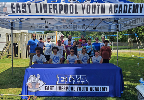 East Liverpool Youth Academy group posing outdoors under a blue canopy with a branded table in front