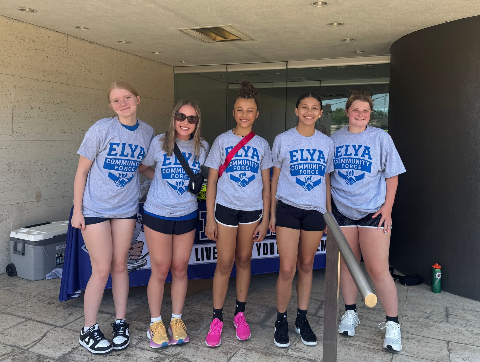 Five athletes in gray ELYA t-shirts pose under a covered walkway, smiling at the camera.