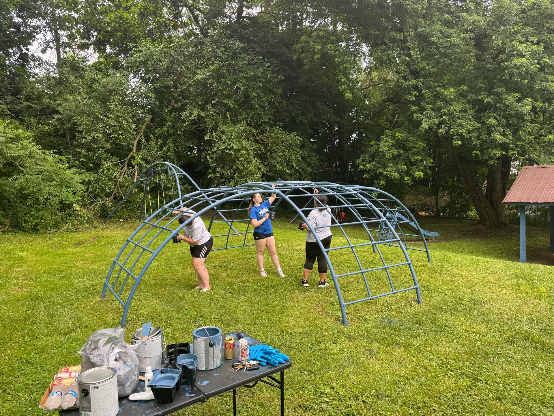 Three people painting a blue dome climbing frame on a grassy lawn