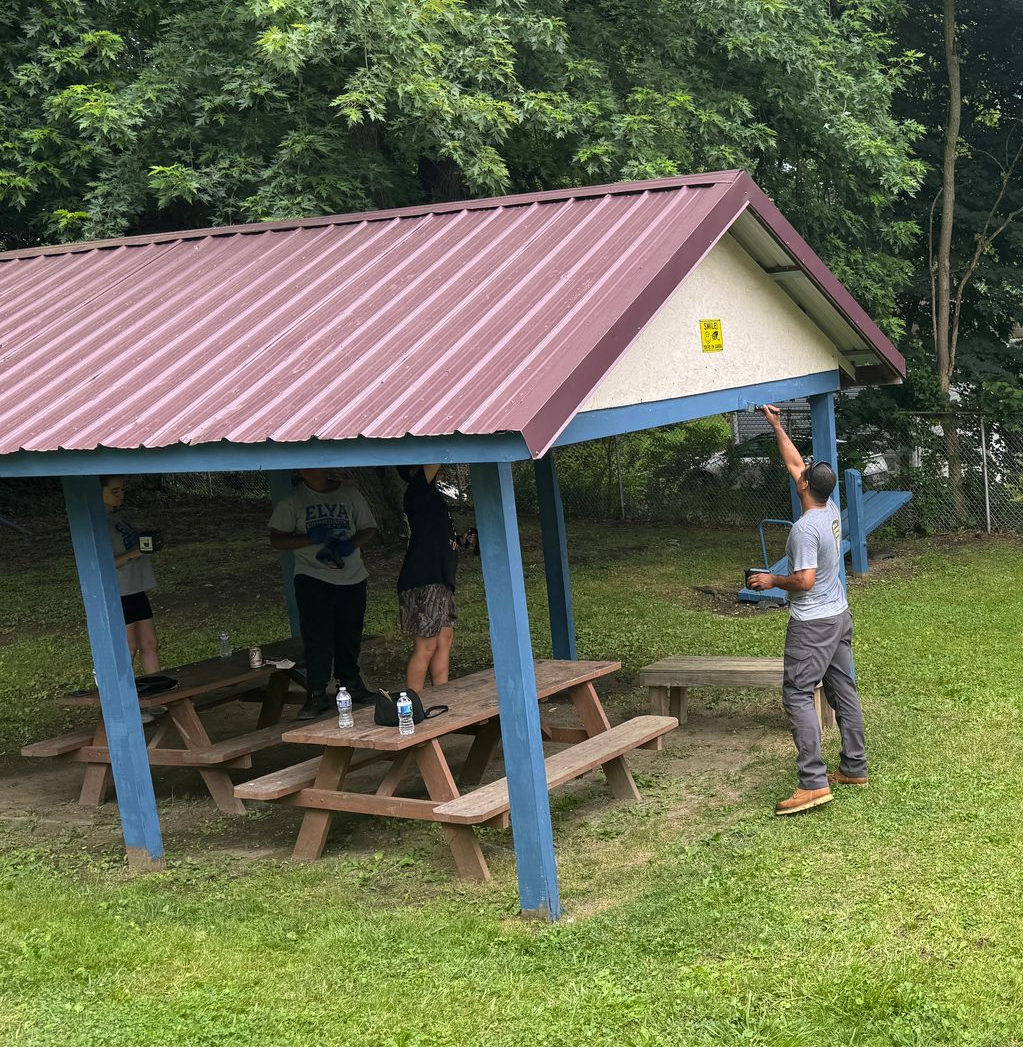 Covered picnic shelter with people, grassy yard, blue playground bars, and cars by a fence.