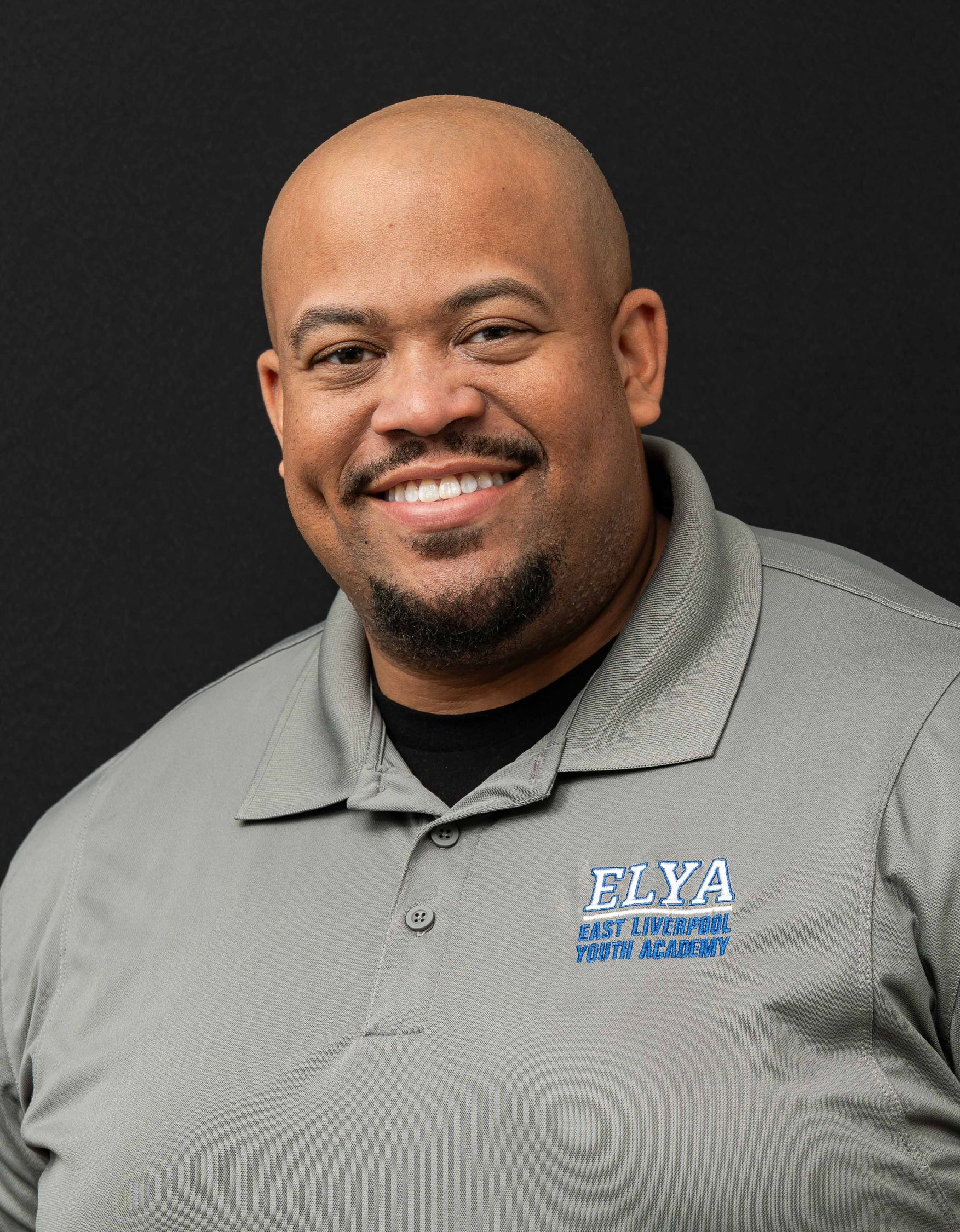 Smiling person in a gray collared shirt with an ELYA logo against a dark background