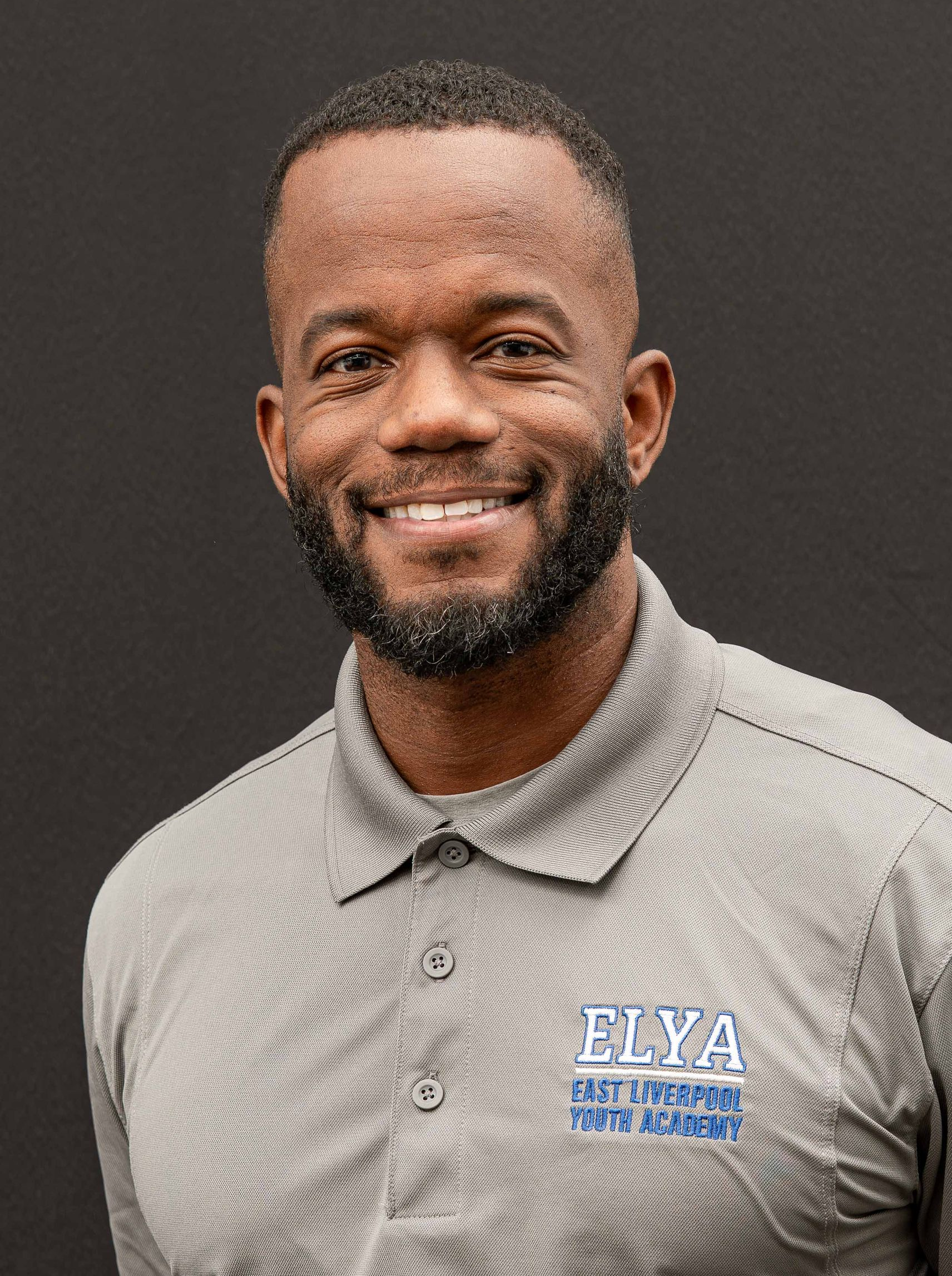 Smiling person in a gray collared shirt with an ELYA logo against a dark background