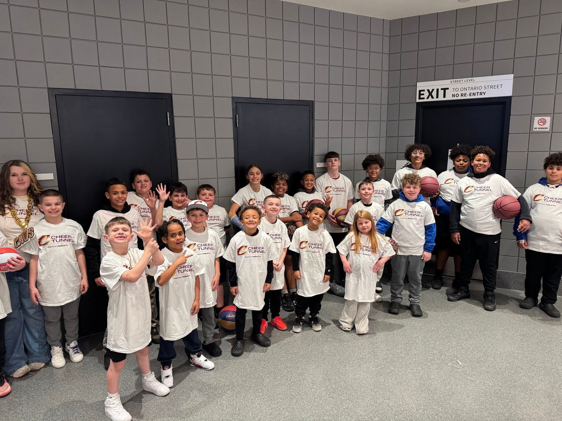 Group of children in white T-shirts posing in a hallway with gray walls and black doors