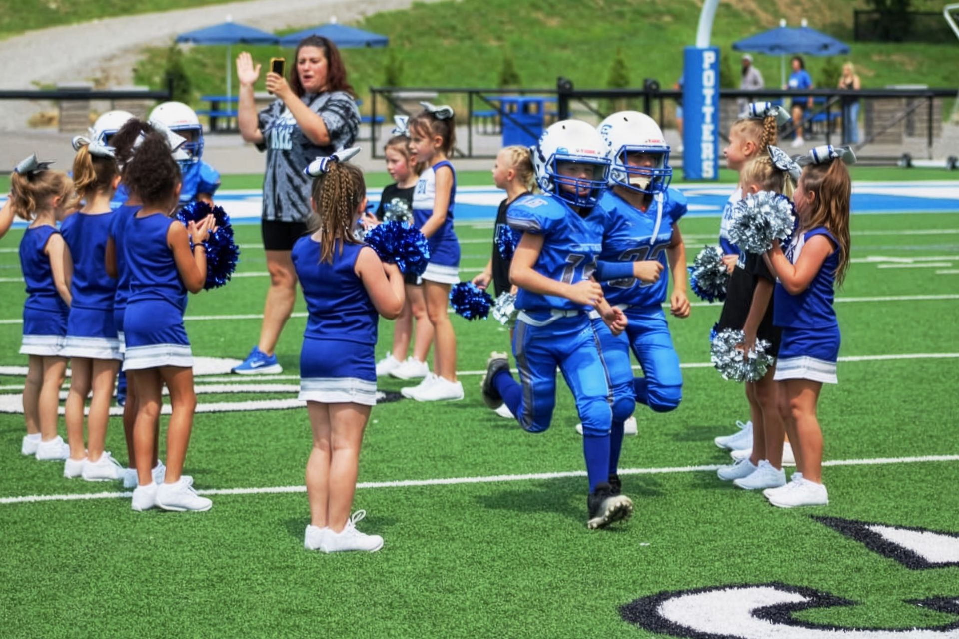 Youth football player runs between blue-clad cheerleaders on a green field during a game celebration