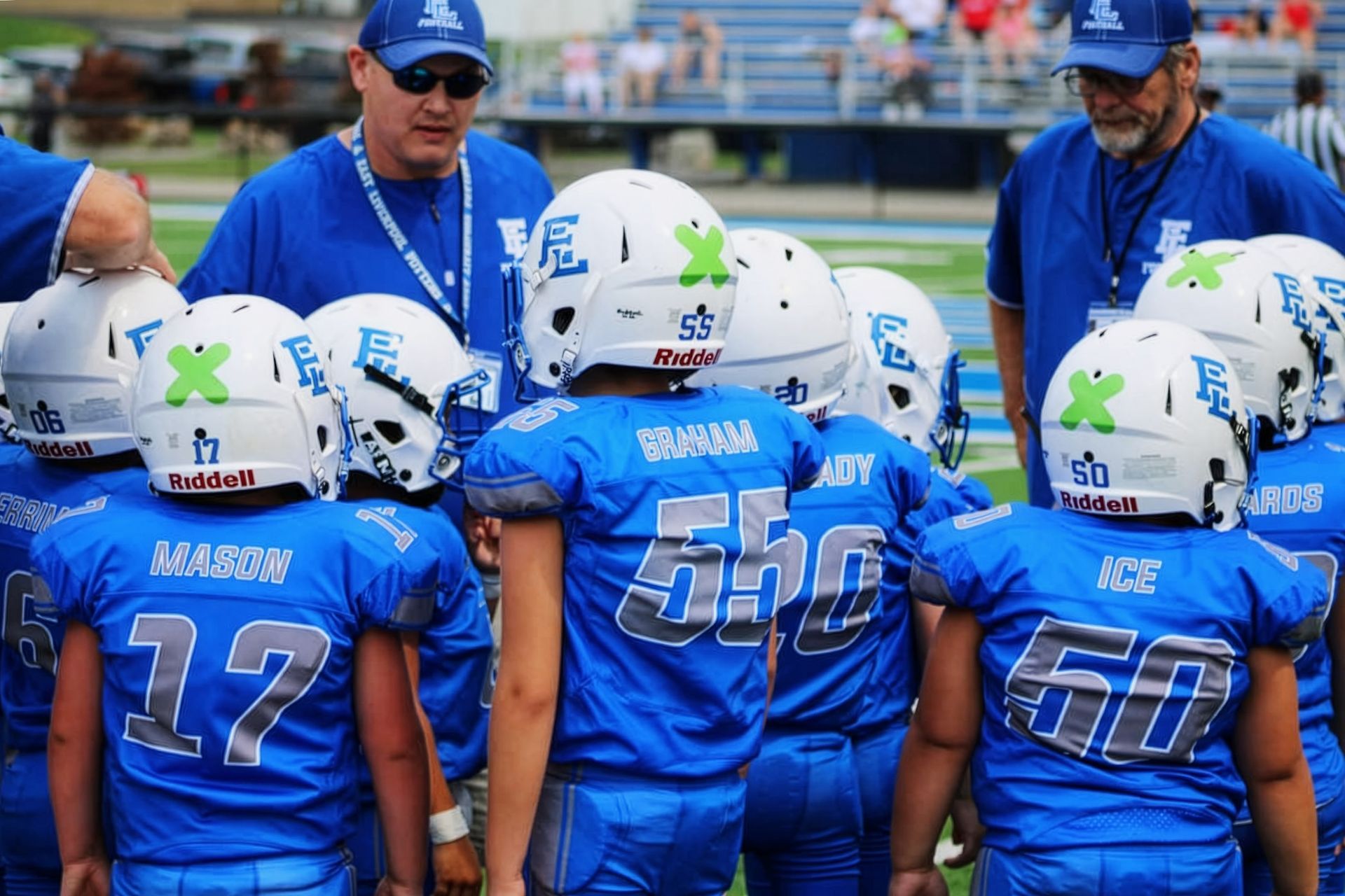 Youth football team huddles with coaches on the field, wearing blue uniforms and white helmets.