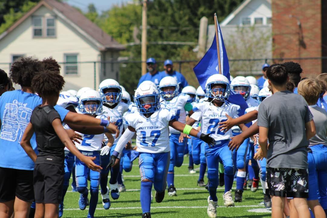 Youth football players in blue uniforms run onto a grassy field between cheering teammates and spectators.