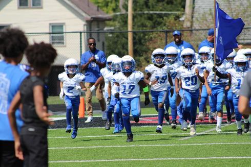 Youth football team in blue uniforms runs onto a grassy field holding flags