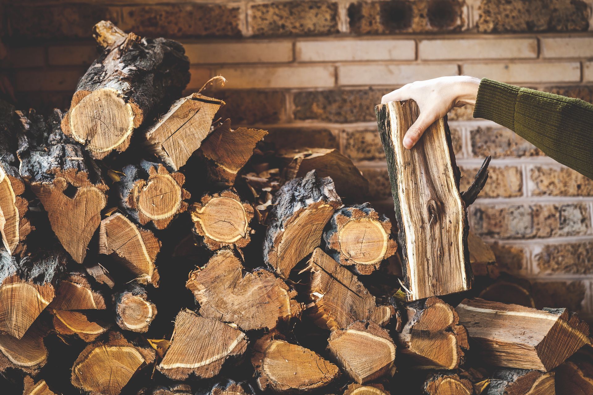 Stacked firewood against a brick wall, with an axe embedded in a log in front.