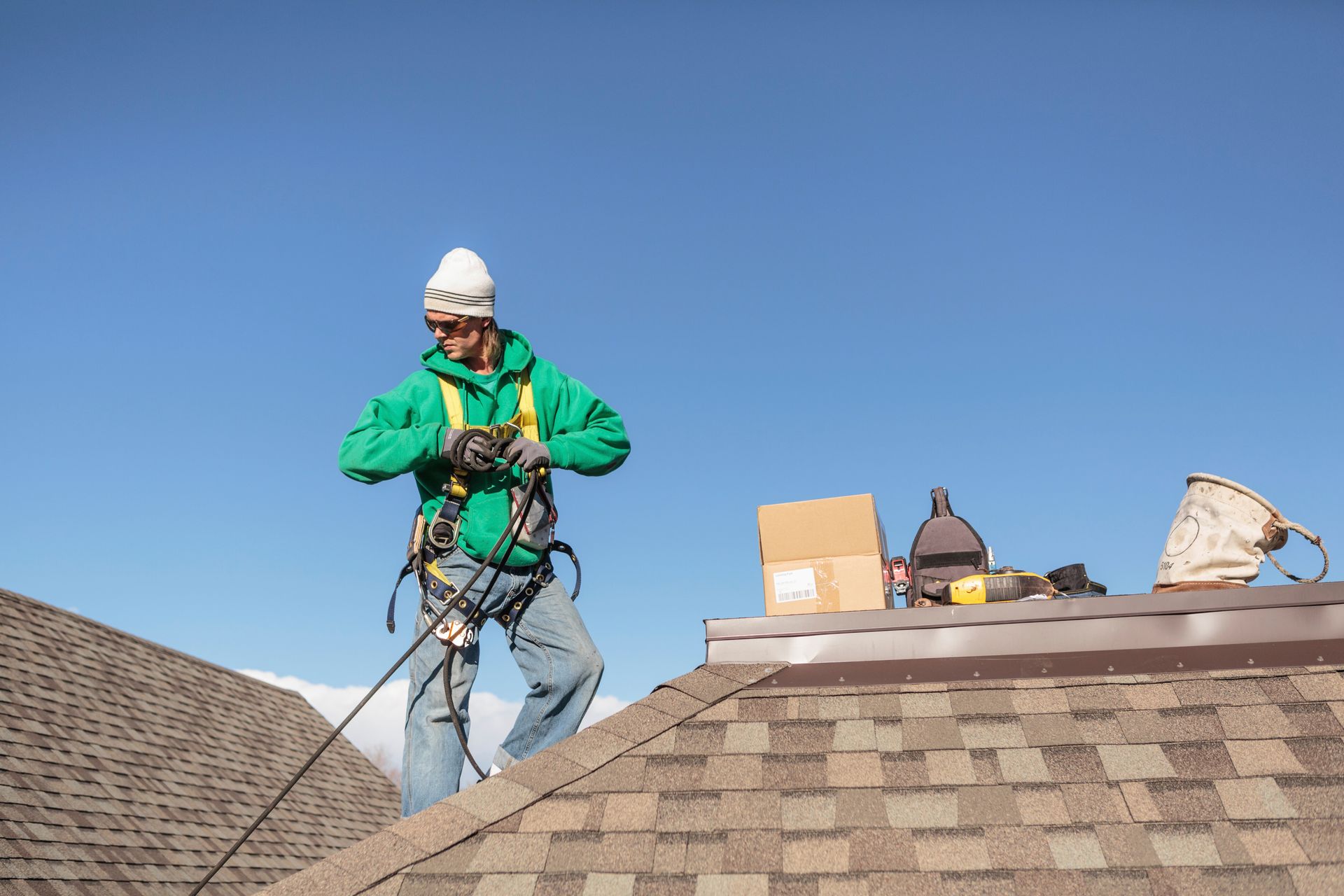 Worker in green jacket and helmet on a rooftop, preparing safety gear beside tools and cardboard boxes.
