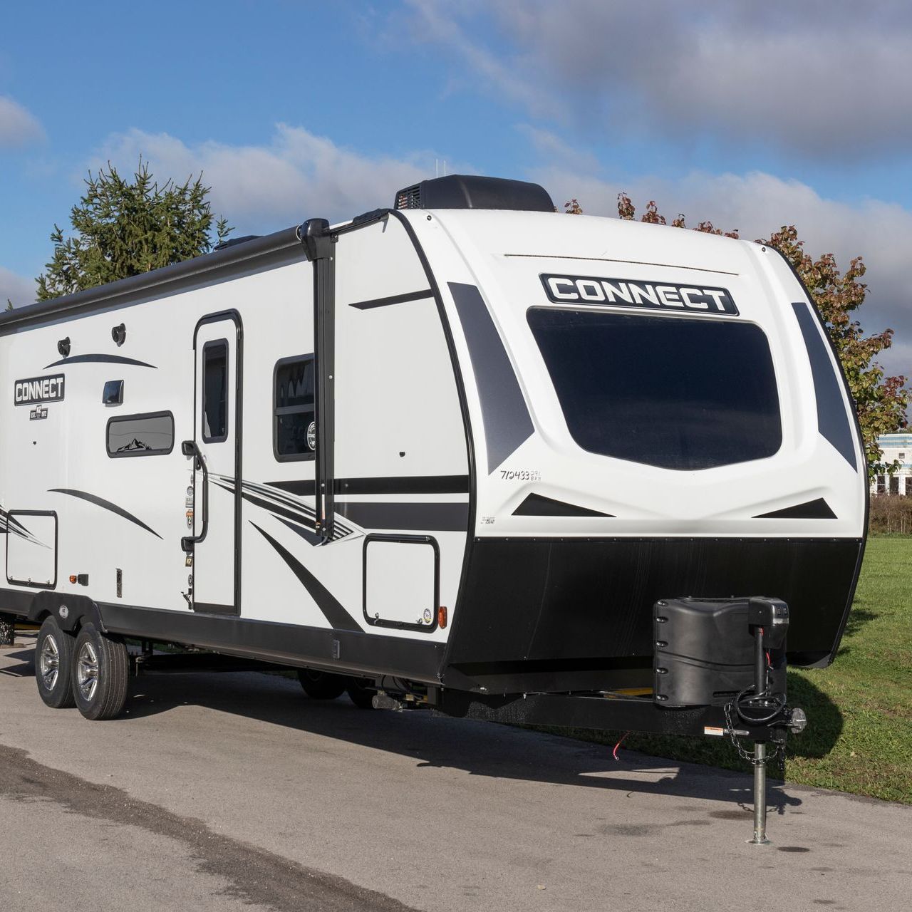 A modern, white and black travel trailer parked on pavement with trees and a blue sky in the background.