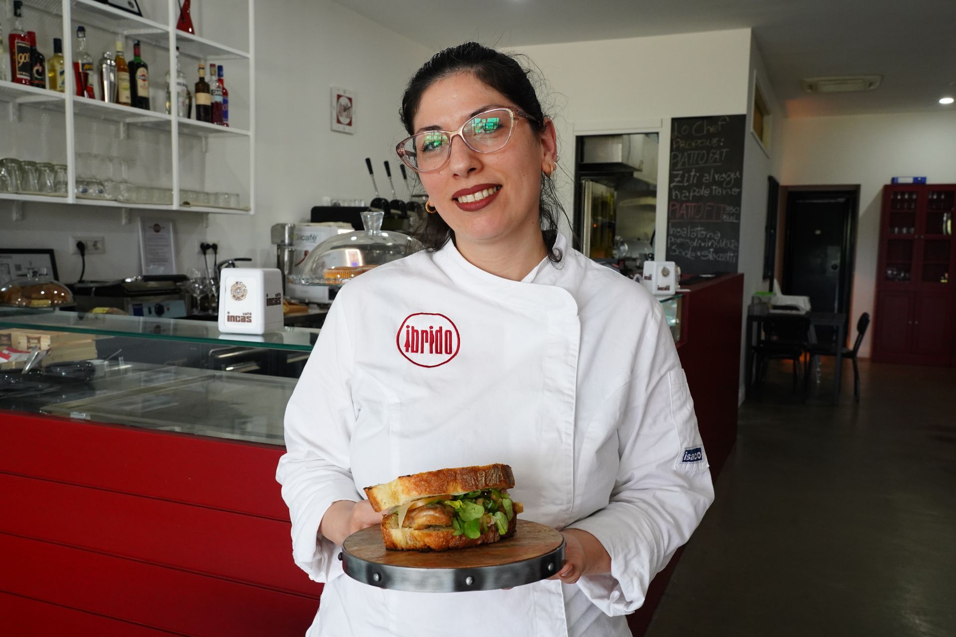 Uno chef in camice bianco tiene in mano un hamburger impiattato nella cucina di un ristorante.
