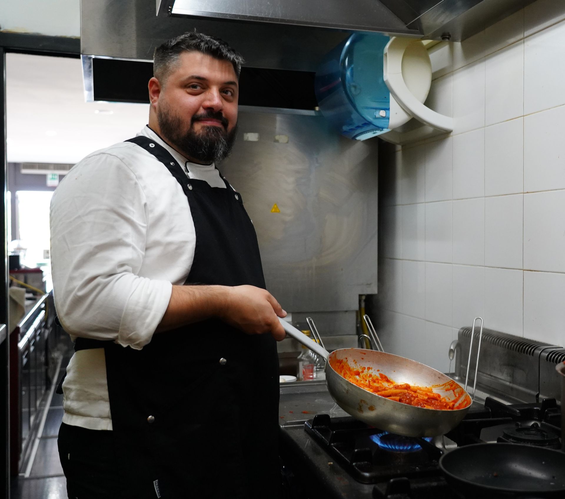 Uno chef in camicia bianca e grembiule nero cucina la pasta sui fornelli in una cucina piastrellata.