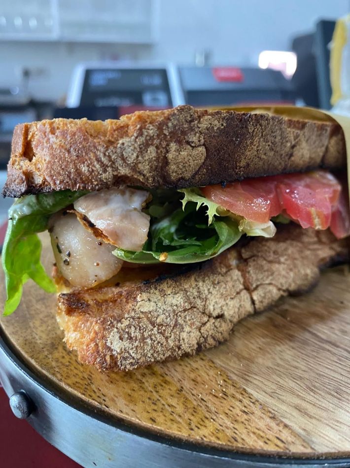 Primo piano di un panino farcito con verdure e pomodoro su un tagliere di legno, con uno sfondo sfocato di una cucina.