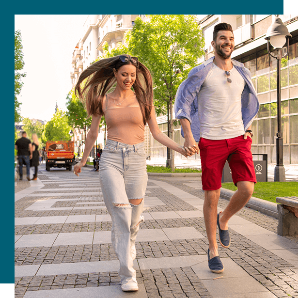 Two people walking on a sunlit city sidewalk, smiling and holding hands, with trees and buildings behind them