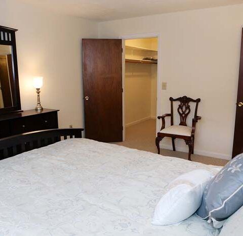 Bedroom with a made bed, chair, dresser, lamp, and open closet door.