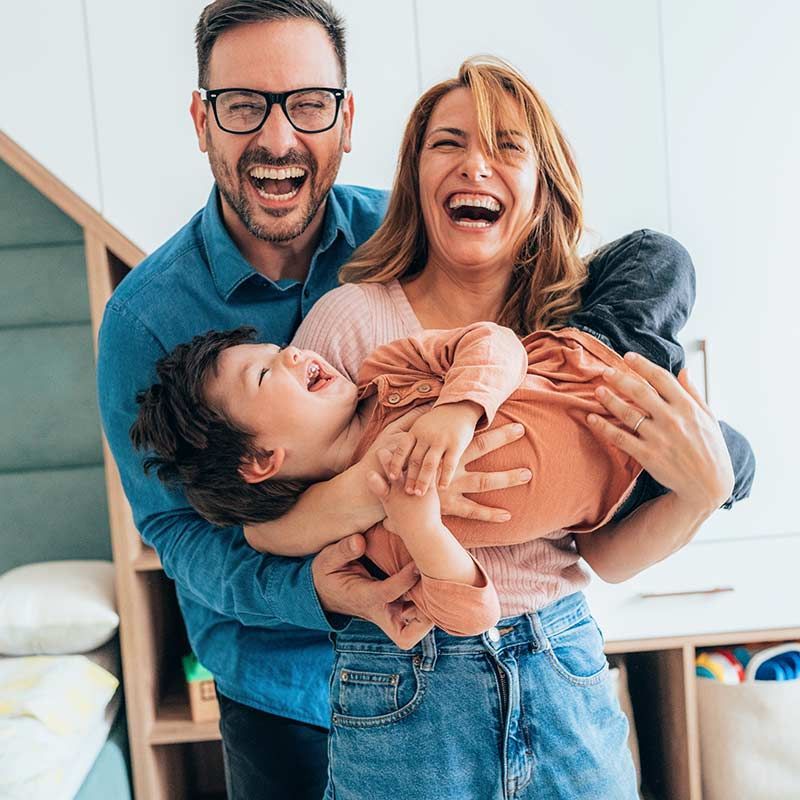 Family of three laughing and hugging indoors, with one leaning into the group and smiling broadly.