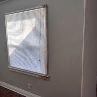 A window with closed white horizontal blinds on a gray wall with white baseboards and trim.