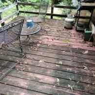 A weathered wooden deck with dark stains, holding a round metal table, chairs, and a propane tank near a railing.
