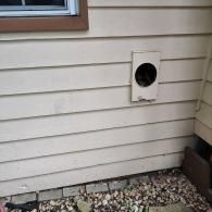 A beige-sided wall with a circular vent opening and a wooden window frame at the top, situated above a gravel ground area.