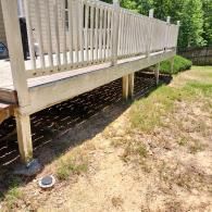 A side view of a raised wooden deck with white railing, supported by posts on a dirt yard with sparse grass.