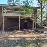 A newly constructed wooden deck with stairs attached to the side of a brick house, with construction debris on the ground.